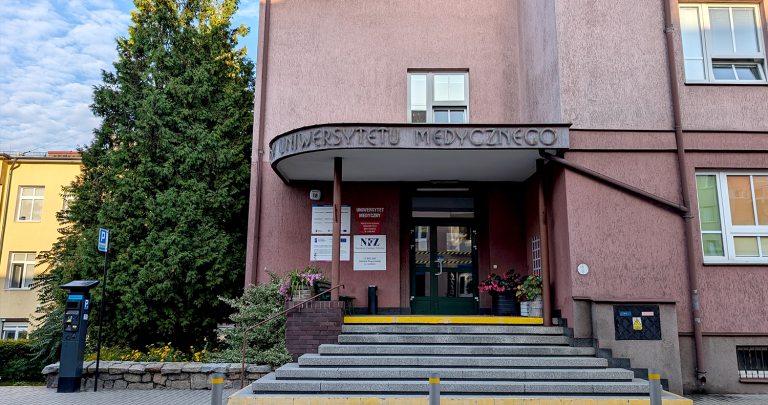 Na zdjęciu znajduje się budynek Biblioteki Głównej. Widoczne są drzwi wejściowe, do których prowadzą schody. Po lewej stronie budynku wyrastają wysokie tuje, stykając się z ciemnoróżową elewacją.