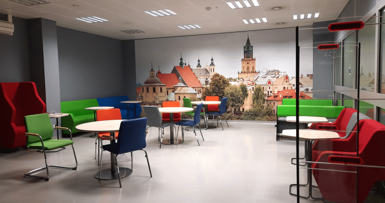 Zdjęcie przedstawia pomieszczenie o ciemnoszarych ścianach i jasnej podłodze. Pokój wypełniają symetrycznie ustawione stoliki oraz krzesła w kolorach zielonym, niebieskim oraz czerwonym. W tle, na jednej ze ścian, znajduje się fototapeta z krajobrazem Lublina.